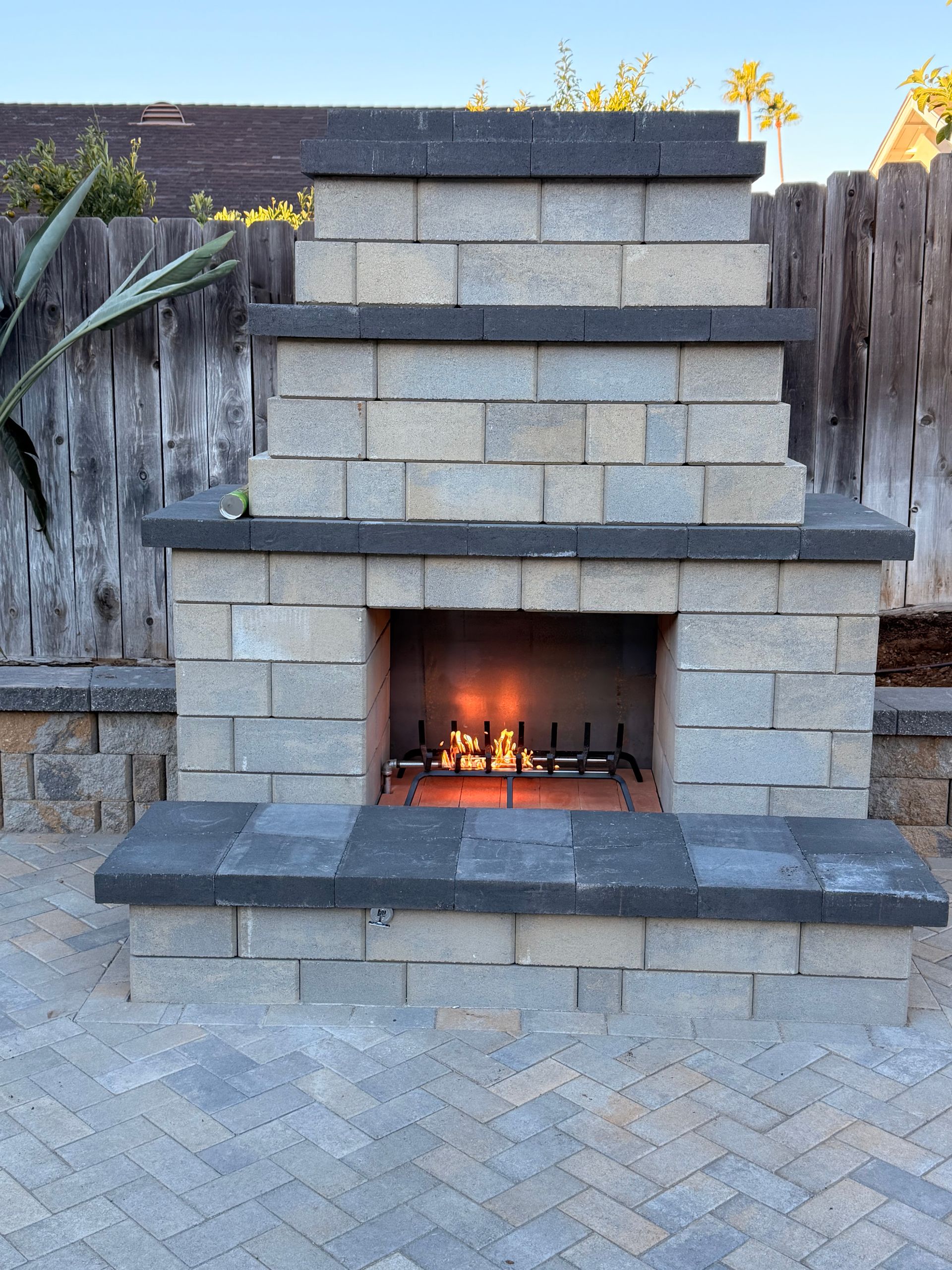 Outdoor fireplace made of light-colored stone blocks with a burning fire, set on a brick patio in front of a wooden fence.