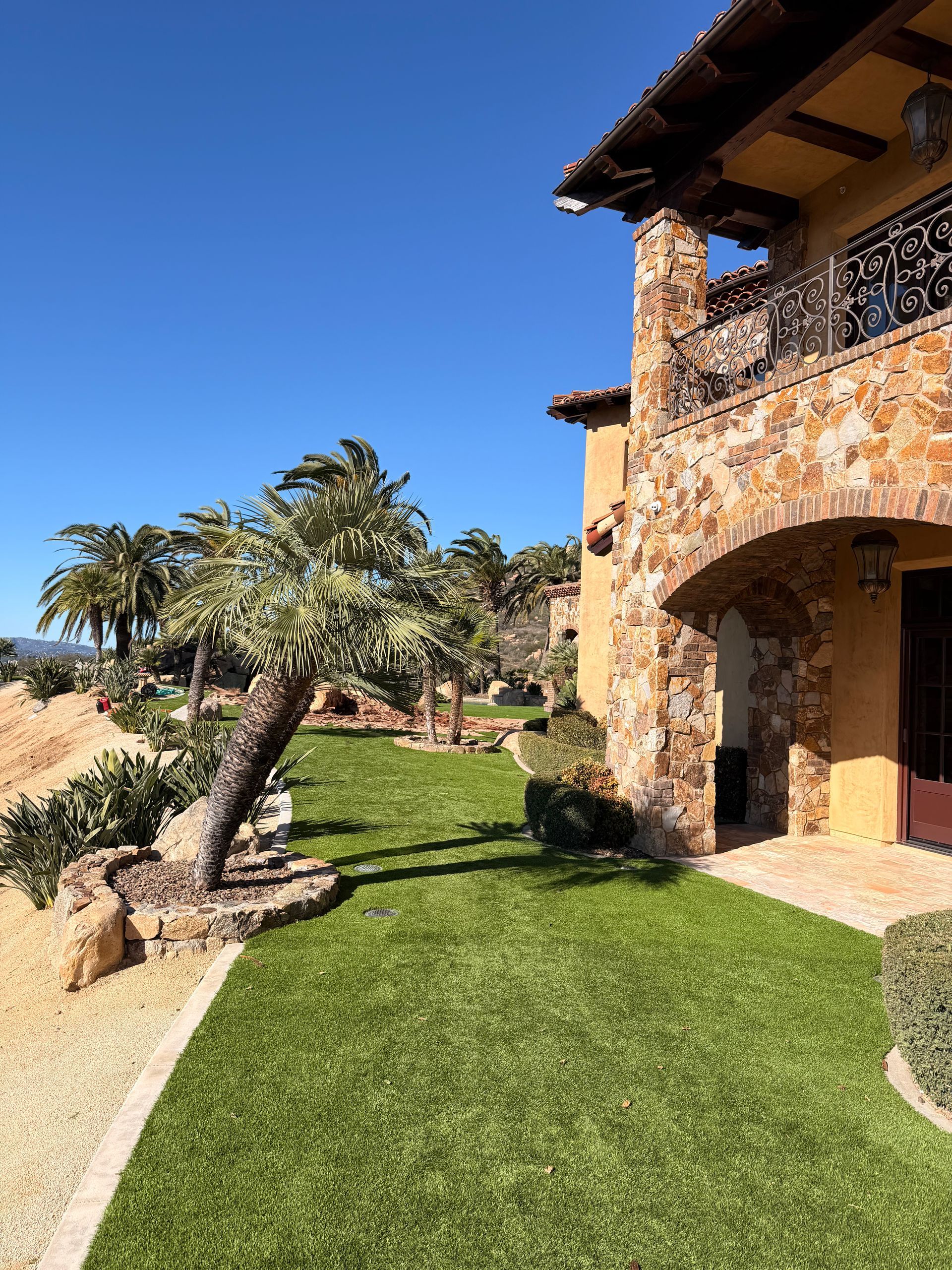 A stone building with a balcony and an arched patio overlooks a green lawn, a leaning tree, and a clear blue sky.