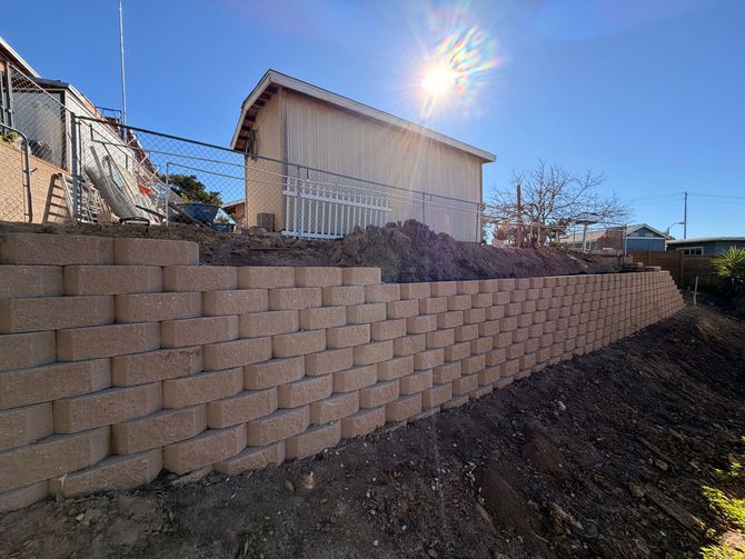 A newly constructed tan concrete block retaining wall slopes upward beside a house on a sunny day.
