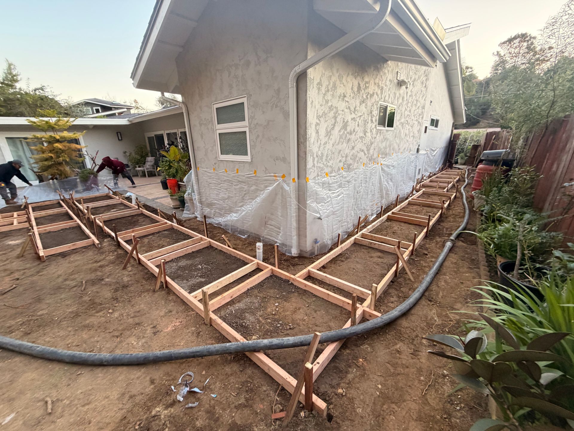 Workers install wooden forms for a concrete sidewalk wrapping around the side of a house at dusk.