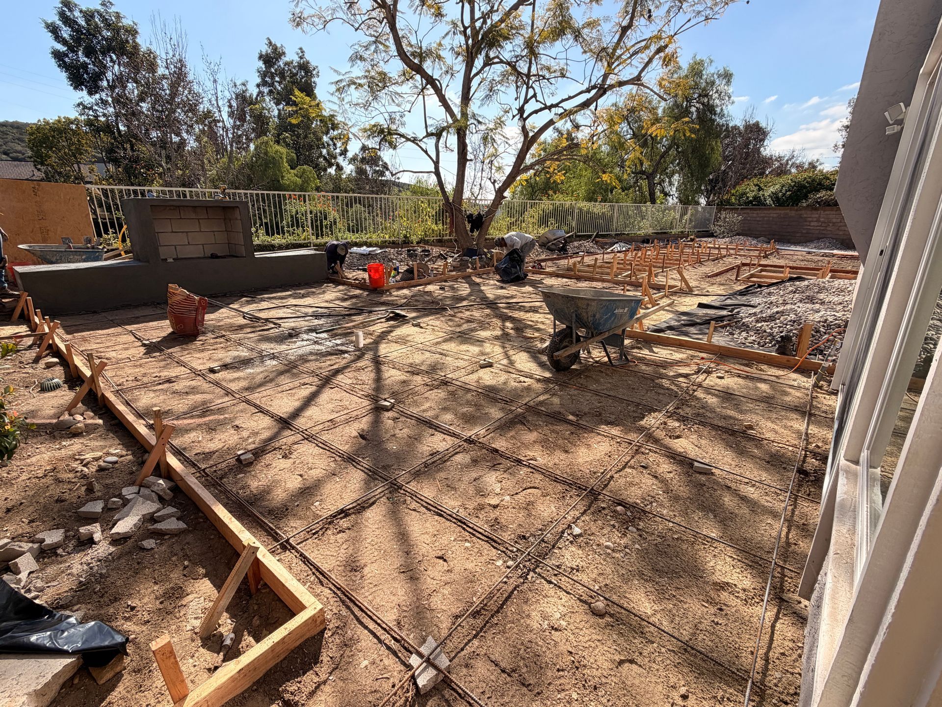 A backyard construction site featuring wooden forms and steel rebar grids laid out on dirt for a concrete patio pour.