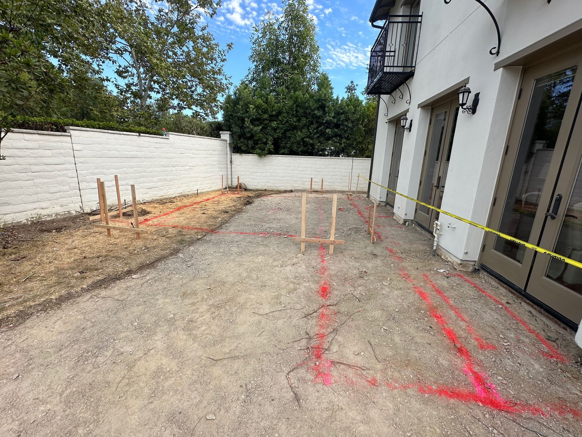 A backyard patio area under construction, featuring wooden stakes, red spray-paint markings on gravel, and safety tape.