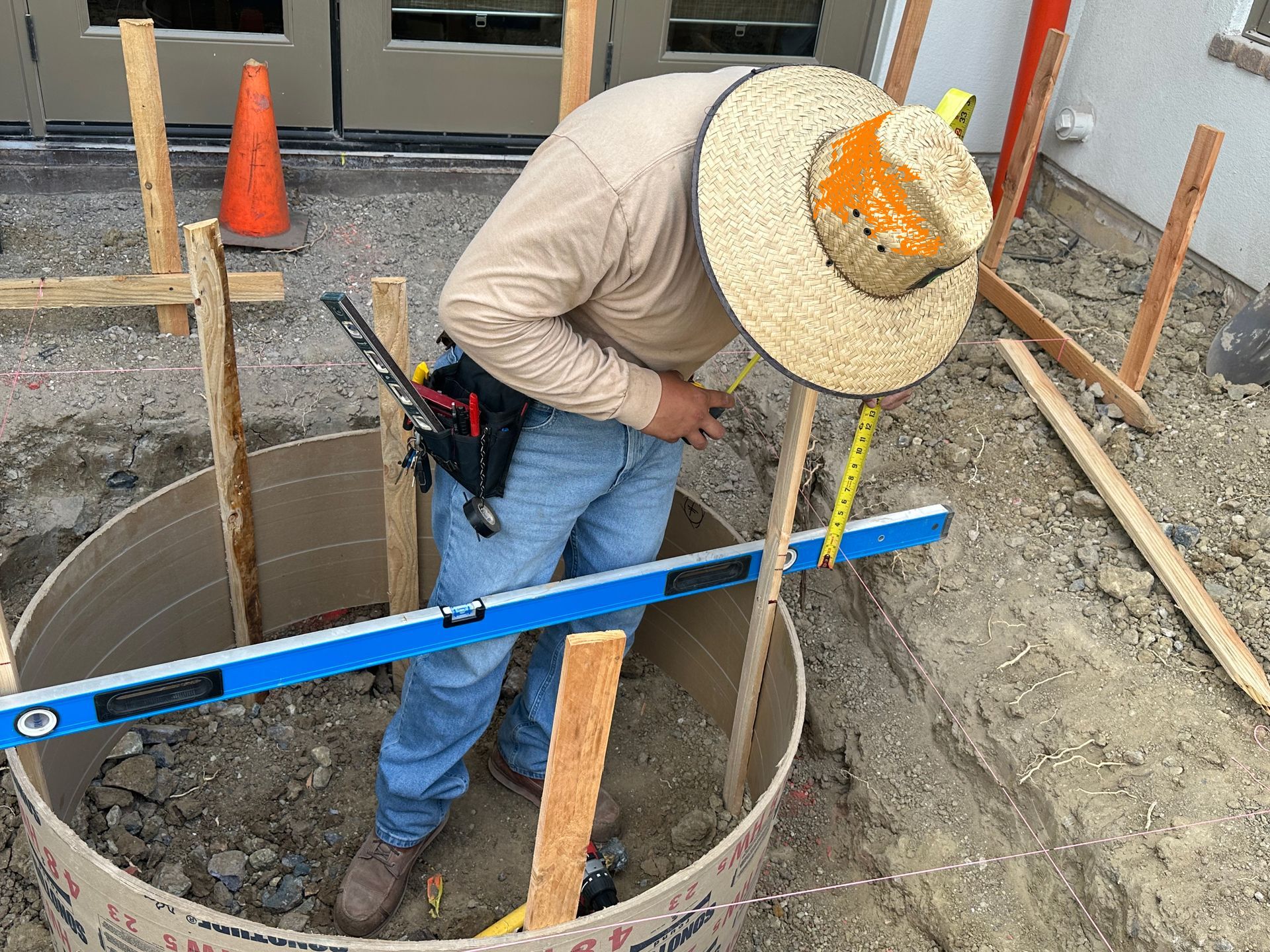A construction worker in a straw hat uses a blue level to measure a circular concrete form outdoors.