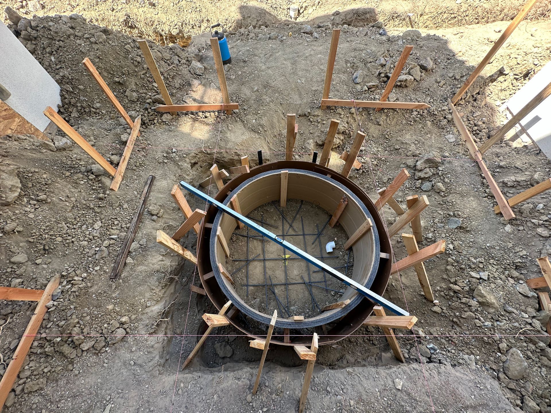 Circular concrete formwork reinforced with wooden braces and a blue level placed on top, set within a dirt construction site.
