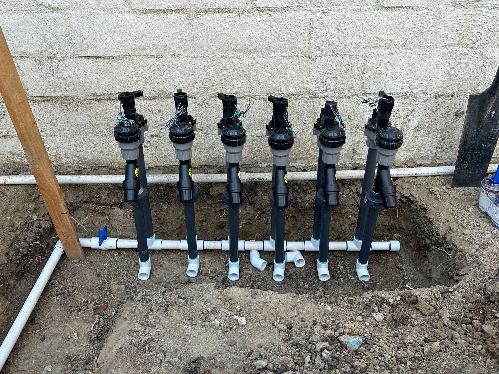 A row of six irrigation solenoid valves connected to a white PVC pipe manifold against a concrete wall.