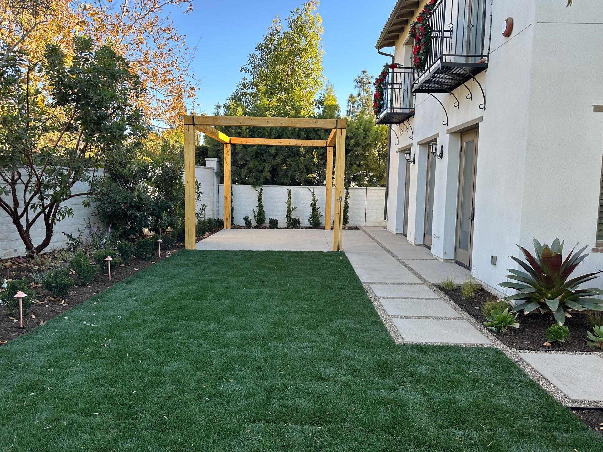 A sunny backyard features a green lawn, a square wooden pergola over a concrete patio, and a white house with a balcony.