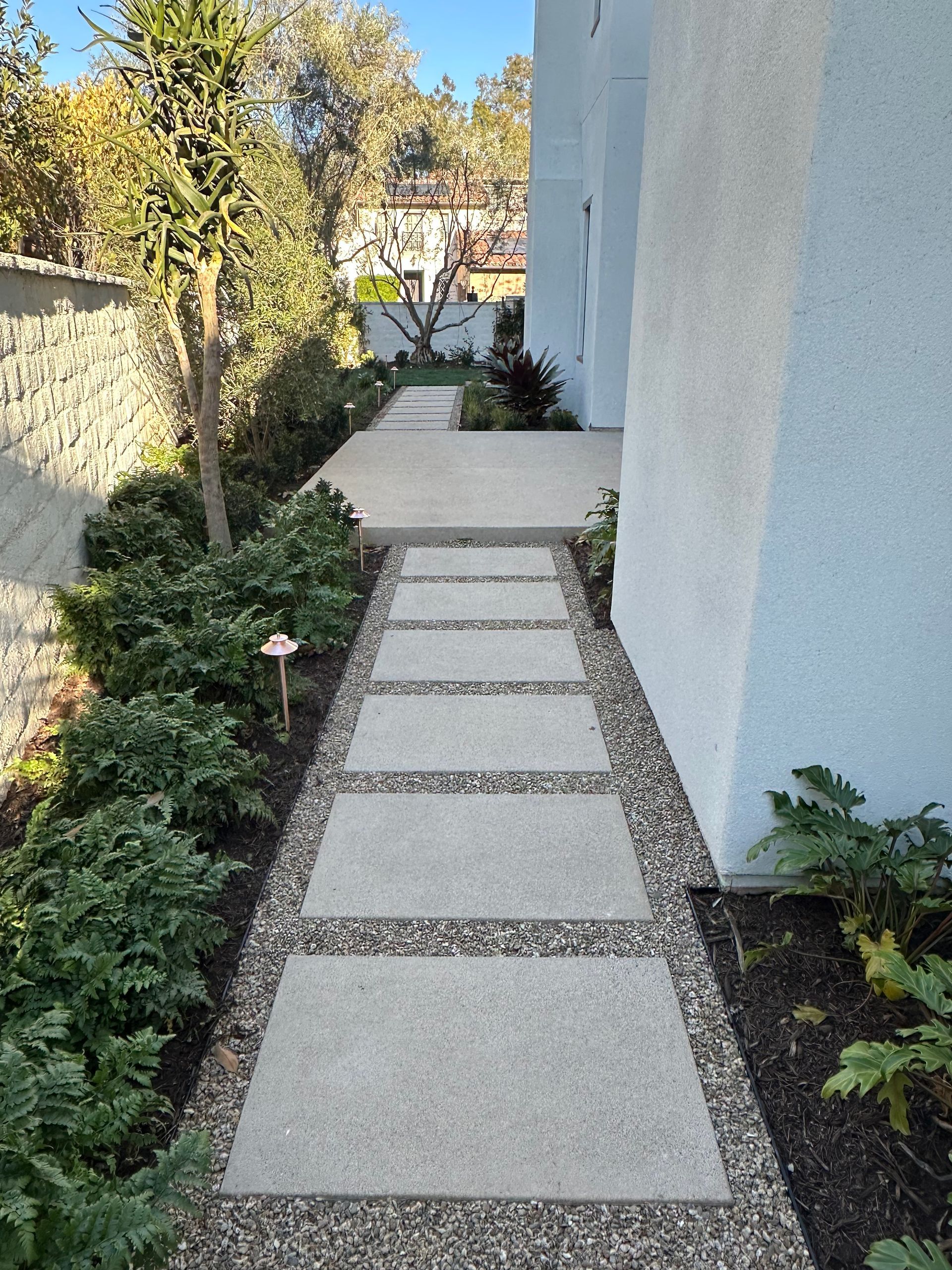 A concrete walkway made of square pavers surrounded by light gray gravel, running between a house wall and garden beds.