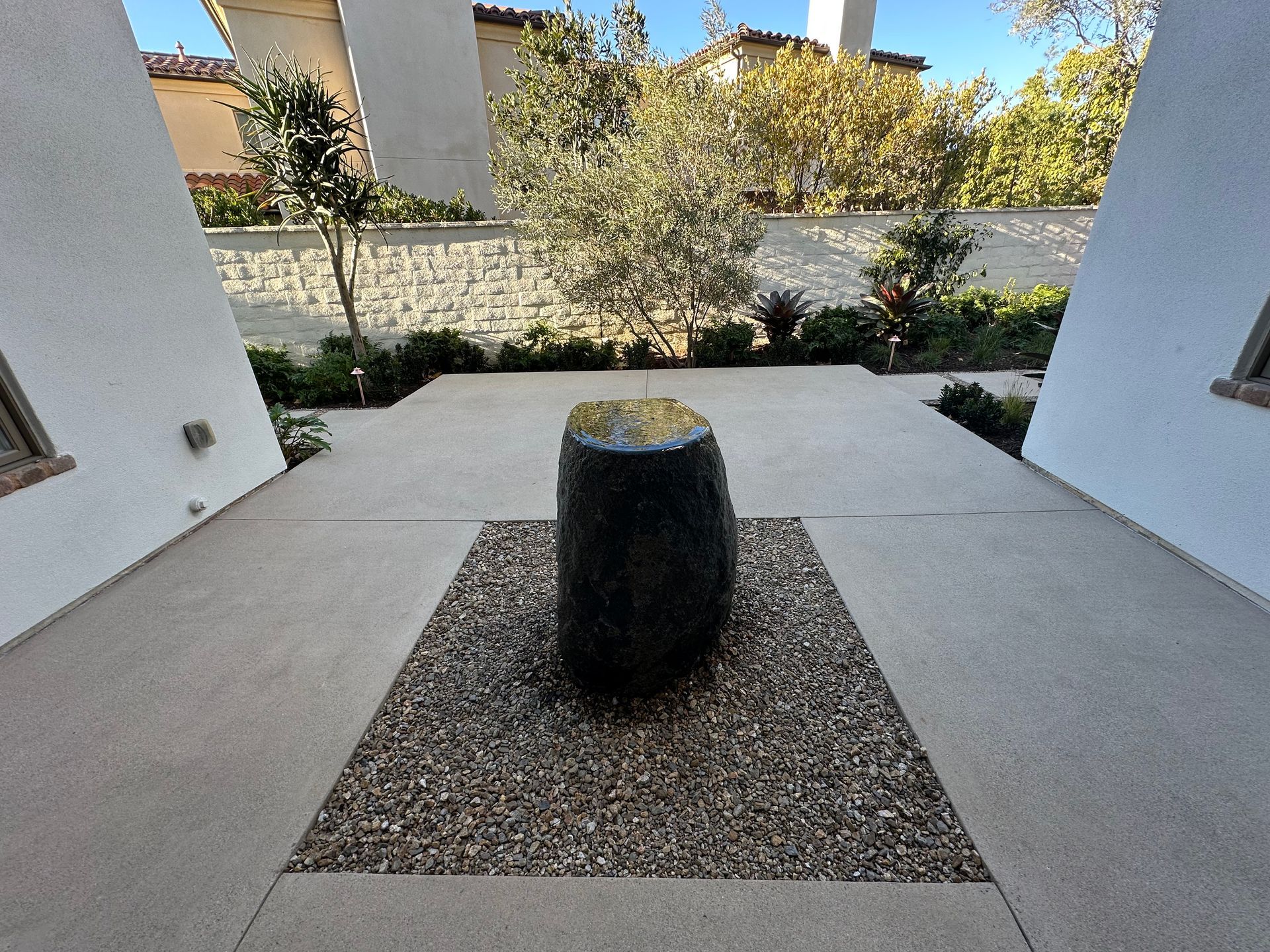 A black, cylindrical stone fountain sits on a square bed of decorative gravel in a light-colored outdoor patio area.