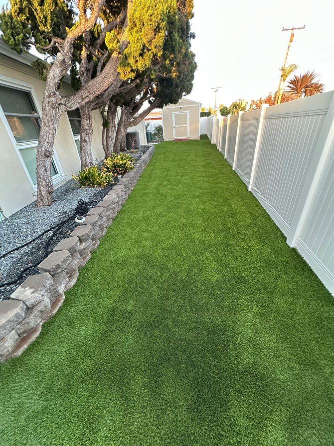 A long, narrow strip of green grass lawn bordered by a stone wall on the left and a white vinyl fence on the right.