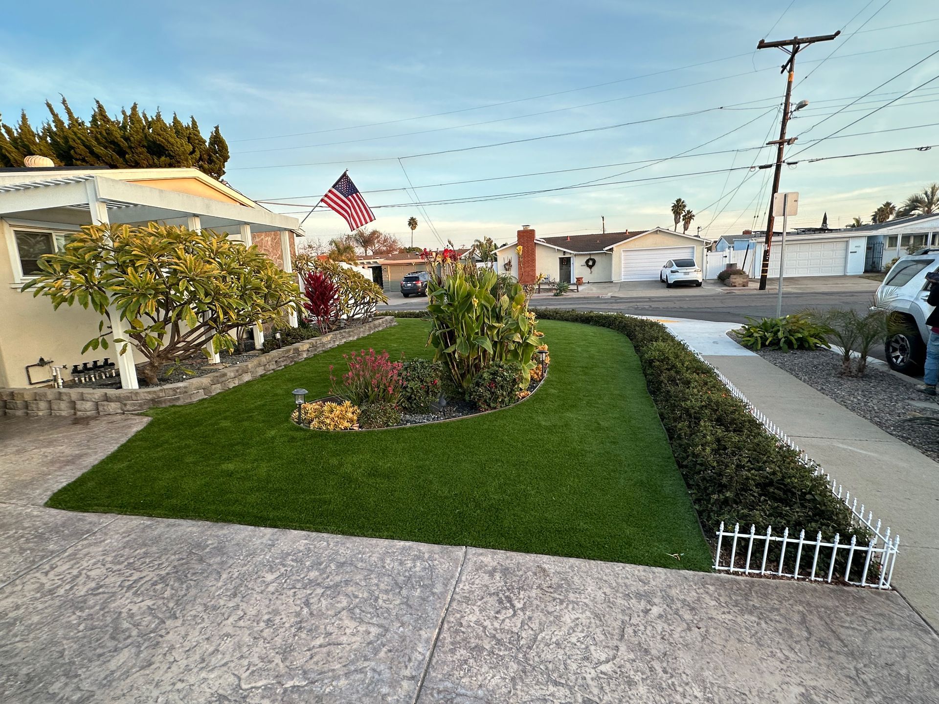 A front yard with a green synthetic lawn, a small garden bed, and a white picket border, set against a residential street.