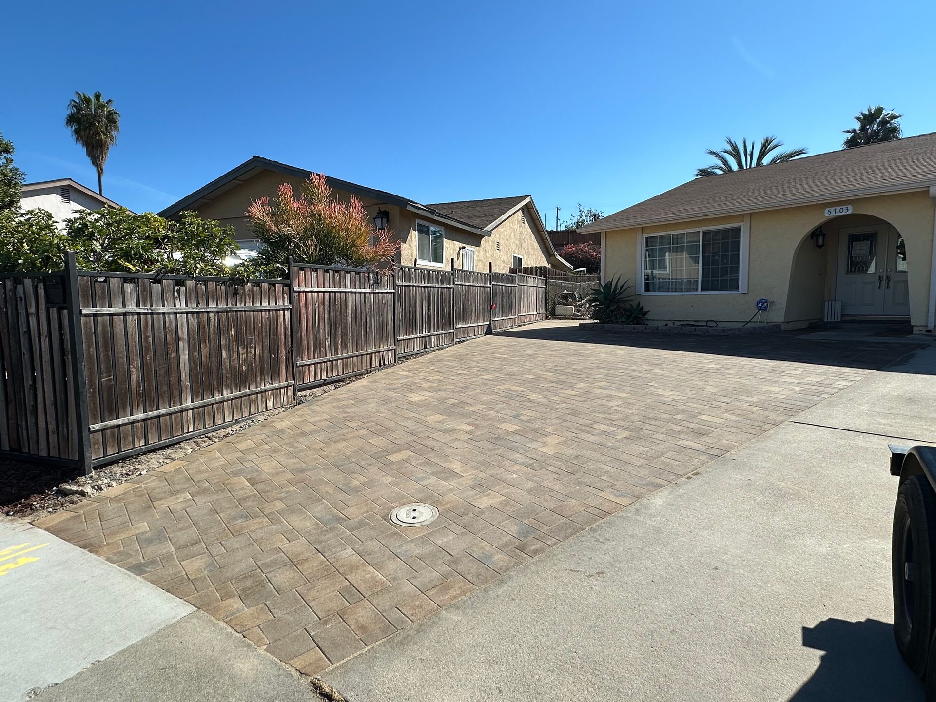 A residential yard with a large paver driveway, a weathered wooden fence, and a light-colored house under a clear blue sky.
