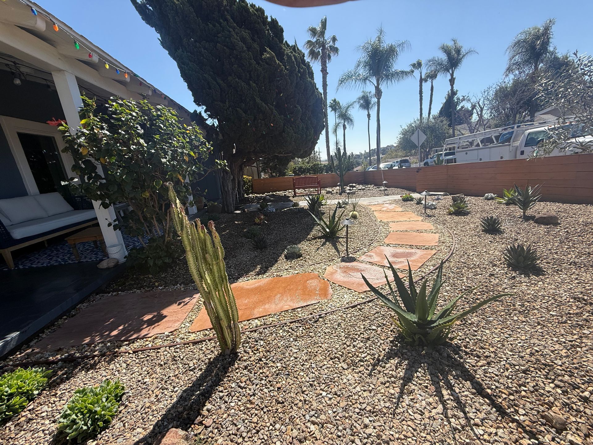 A dry-scaped garden with a stone path leading past aloe plants and a tall cactus, bordered by a home porch and fence.