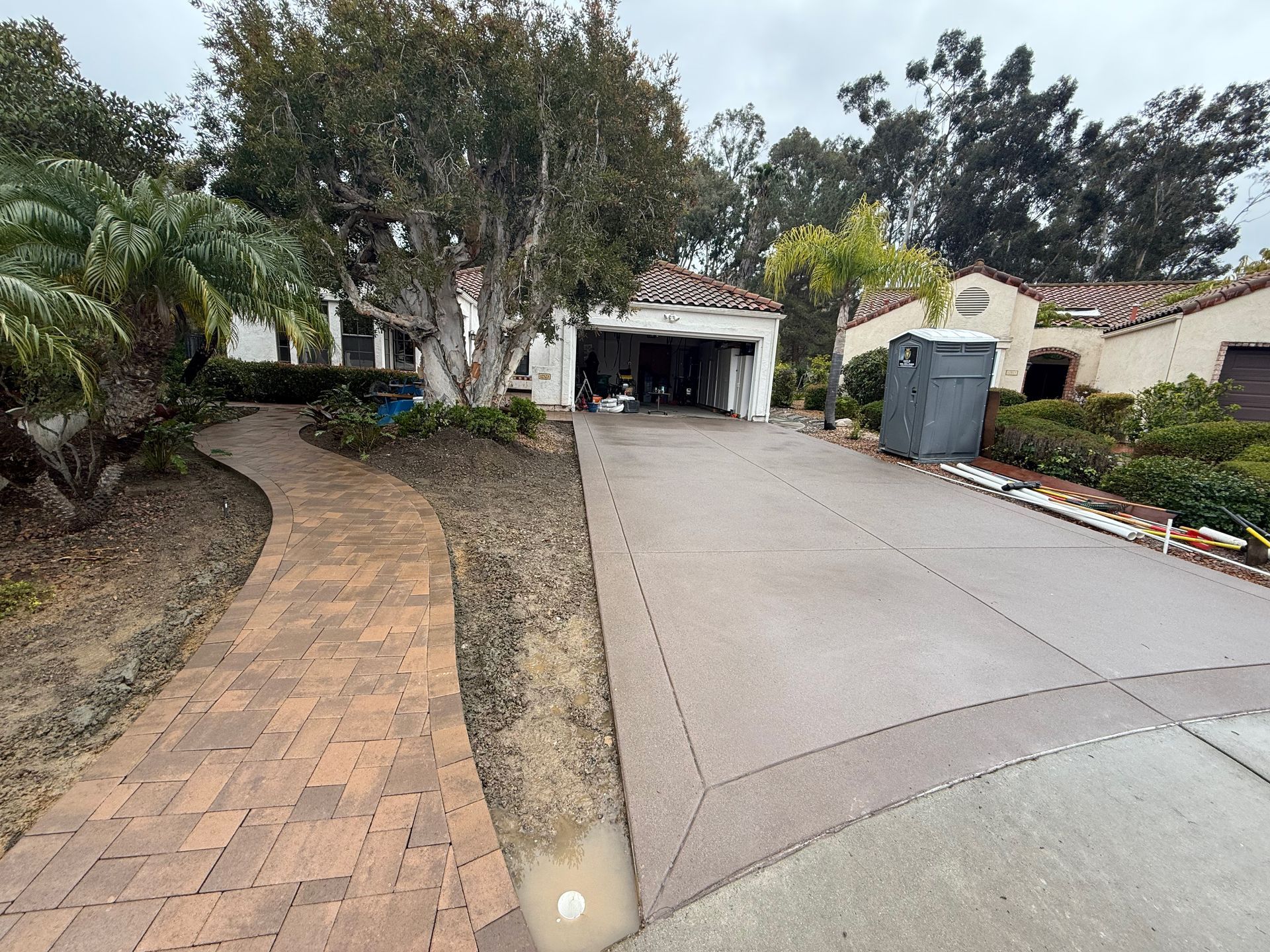 A paved brick walkway leads to a house with a concrete driveway, a large tree, and a portable restroom on a cloudy day.