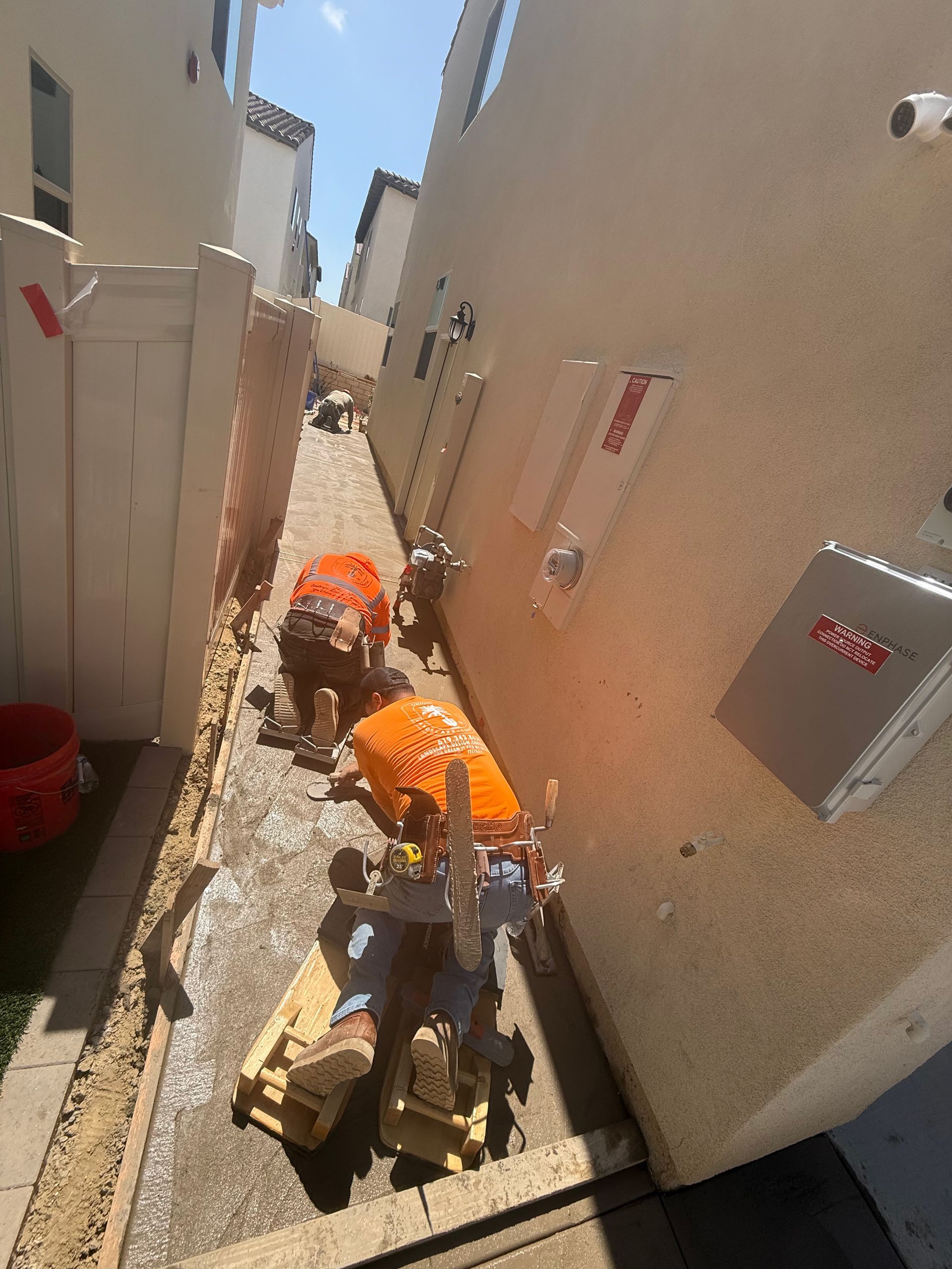 Two workers in orange shirts work on a gravel path in a narrow alley between two residential buildings.
