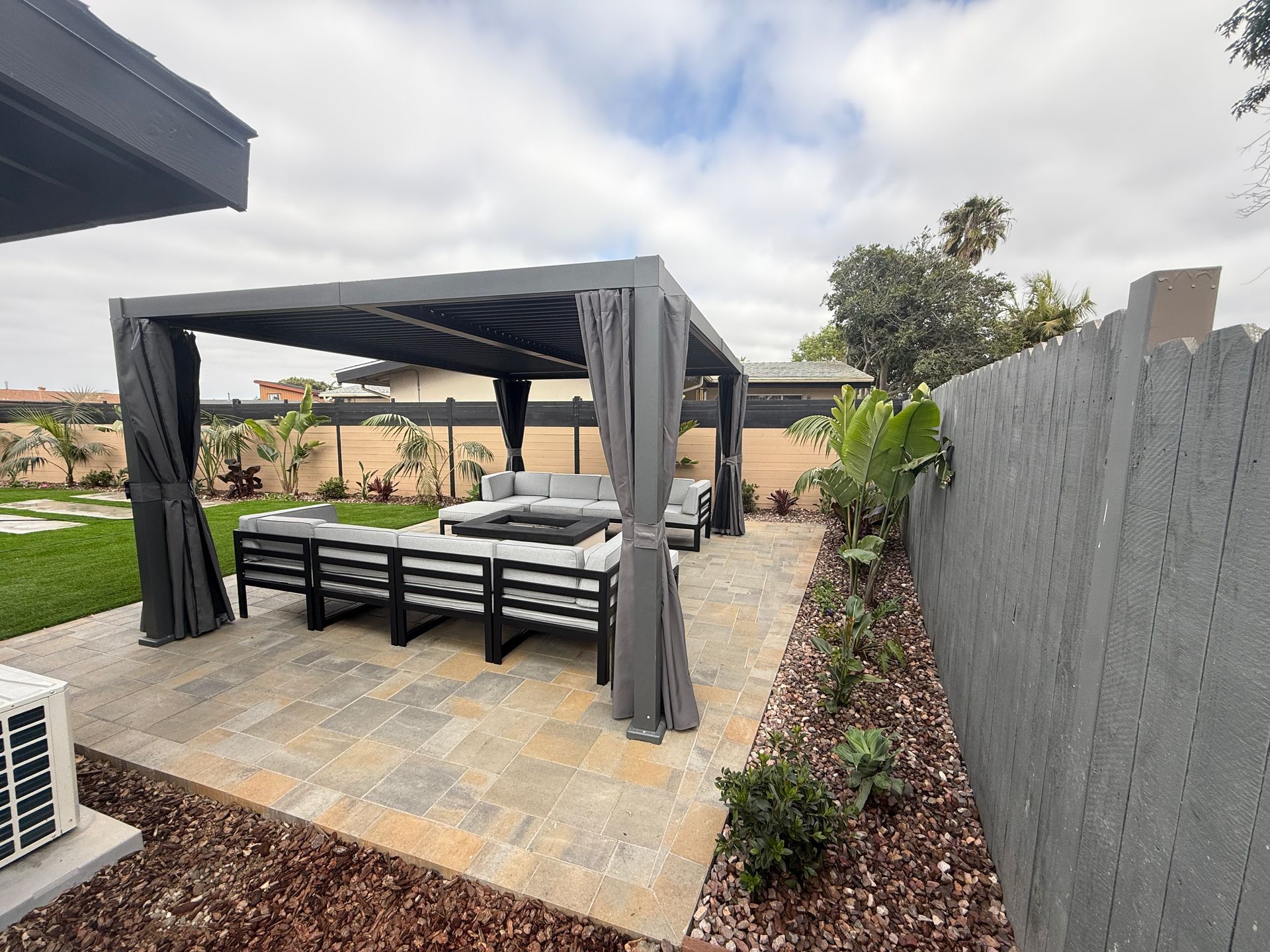 A gray pergola with curtains sits over patio furniture on a stone patio in a backyard with a gray fence and greenery.