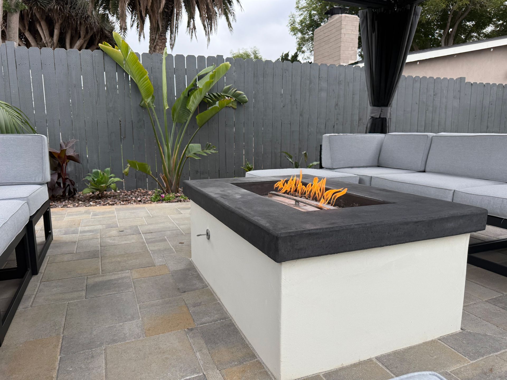 A rectangular fire pit with a black top and white base sits on a stone patio surrounded by grey sectional outdoor seating.