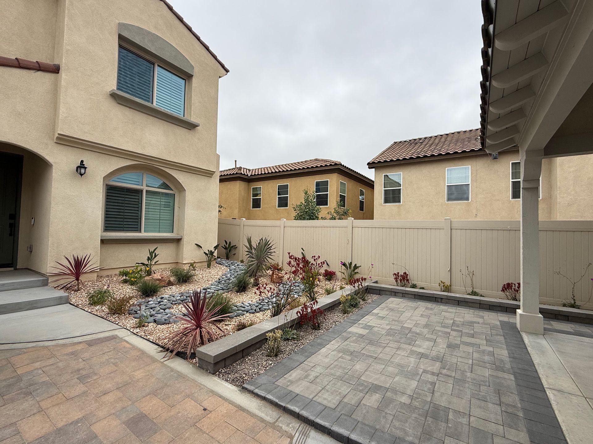 A courtyard area with stone pavers, a xeriscaped rock garden with small shrubs, and a beige stucco house under a gray sky.