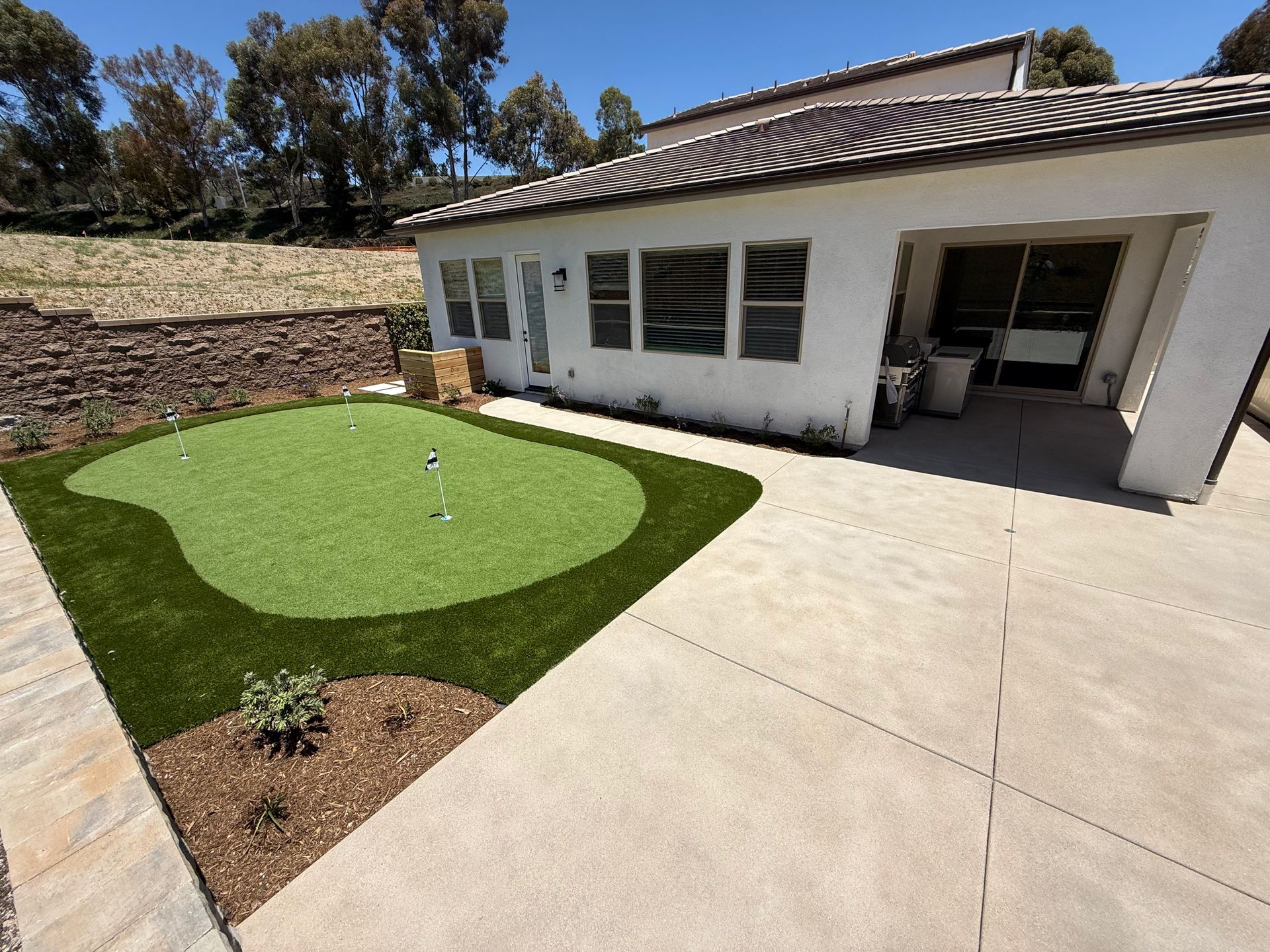 Backyard featuring a synthetic putting green with two cups, a concrete patio, and a stone retaining wall under a blue sky.