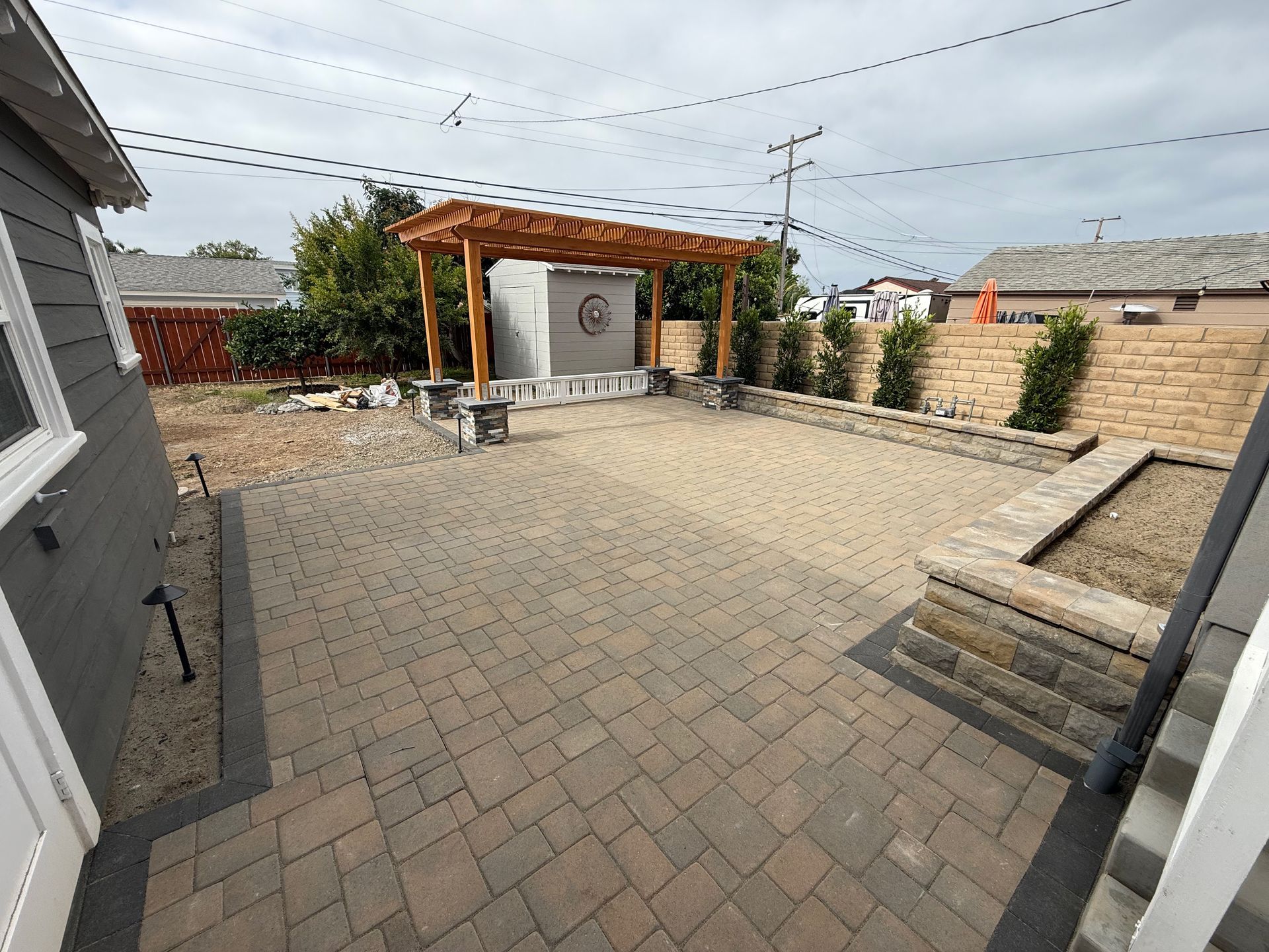 A backyard patio featuring tan paved stones, a wooden pergola, a stone planter wall, and a gray shed under a cloudy sky.