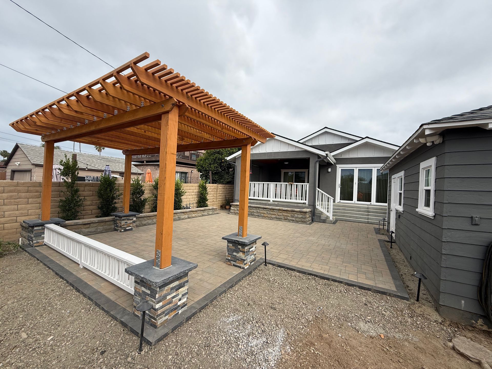 A wooden pergola sits over a paved patio next to a gray house, with stone pillar accents and a white bench.