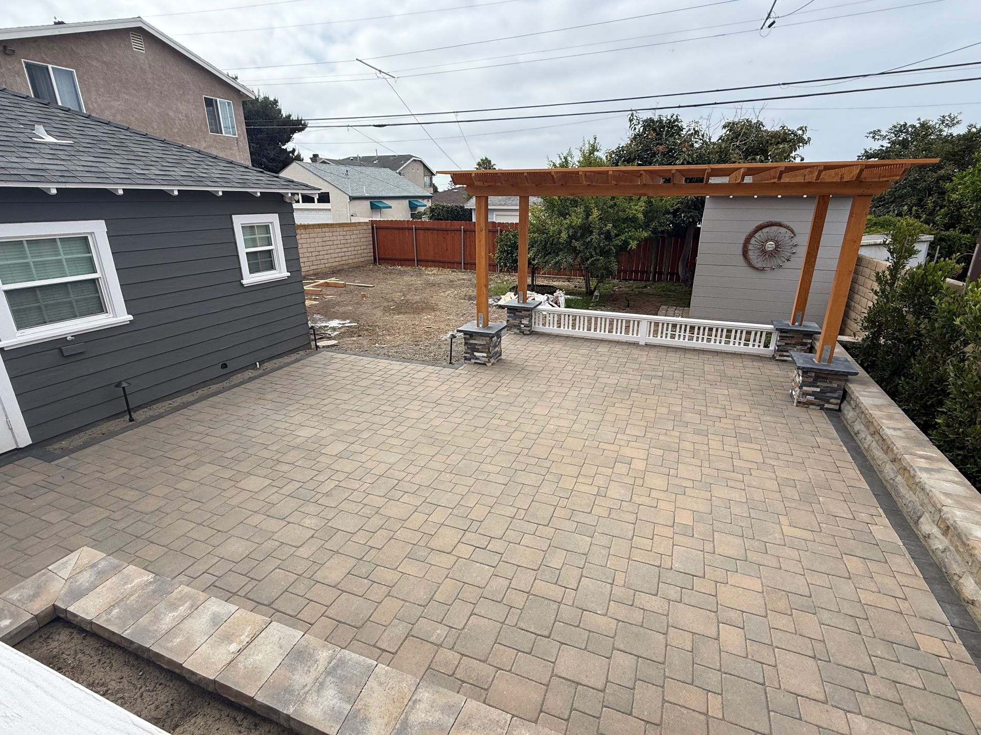 A paved patio area with a wooden pergola, gray siding house, and a small patch of dirt in a residential backyard.