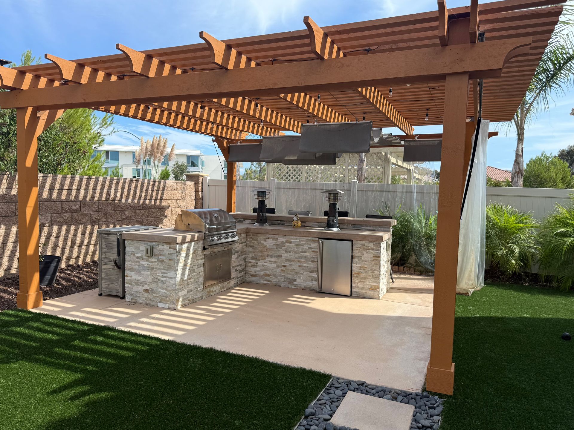 An outdoor kitchen with a stone base and stainless steel appliances under a wooden pergola on a patio next to a lawn.