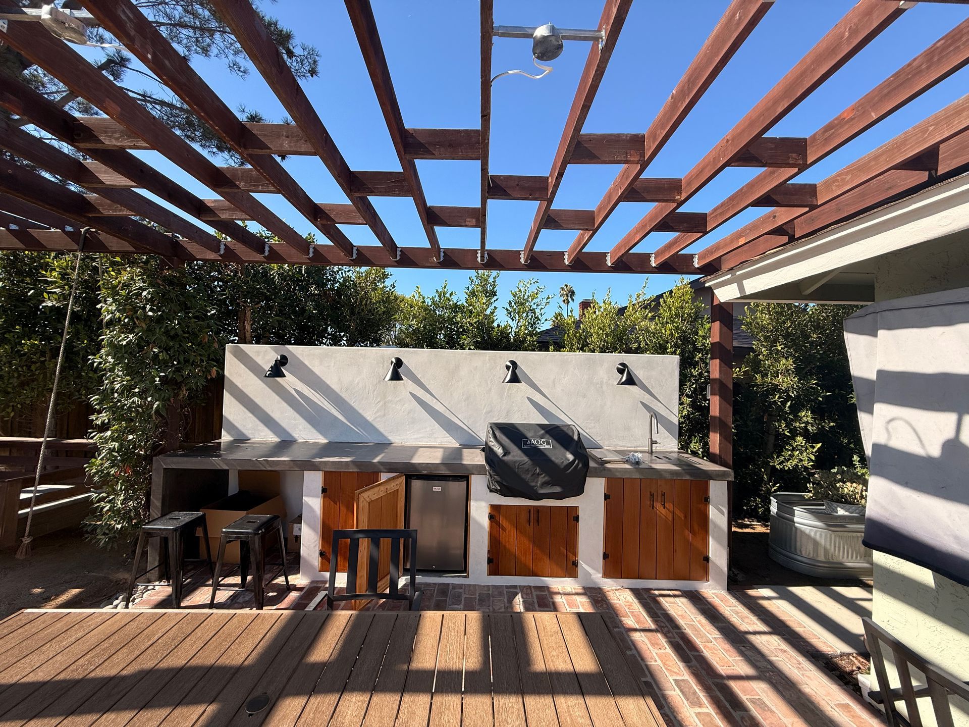 An outdoor kitchen with a grill, wooden cabinets, and a concrete counter under a wooden pergola on a sunny deck.