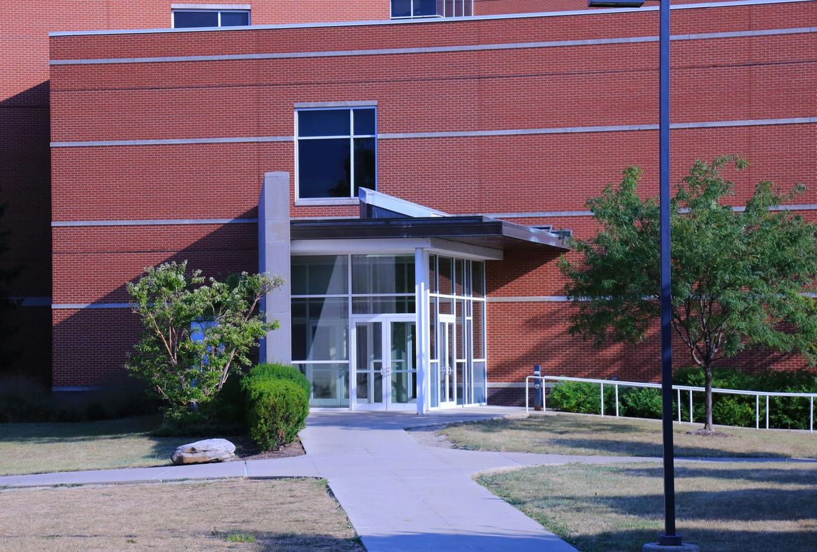 A brick building entrance with a glass-paneled door, a metal awning, a walkway, and small trees on a sunny day.