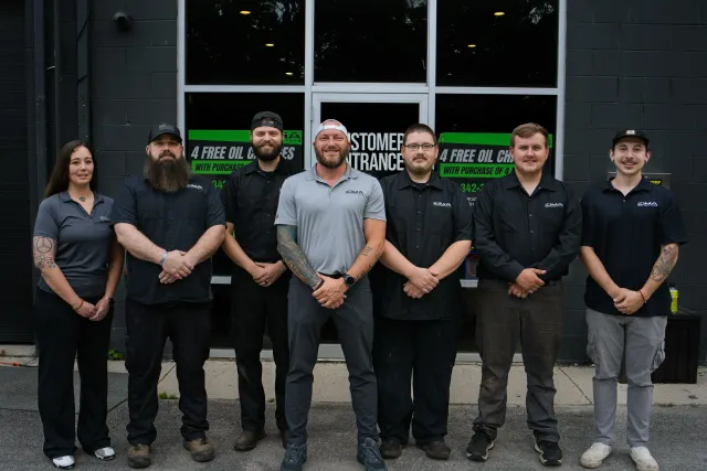 Zima team posing outdoors, holding a sign with a black Zima logo, one seated beside a small fire