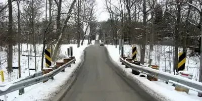 Snowy road crossing a narrow bridge with guardrails and bare trees on both sides