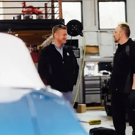 Two men talking in a garage workshop, with a blue car in the foreground.