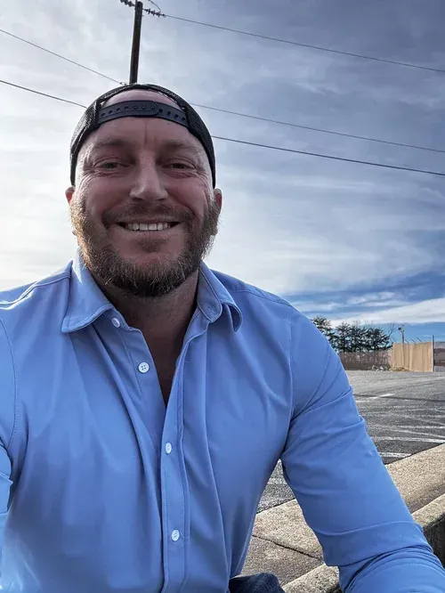 Smiling person in a backward cap and light blue shirt outdoors near a road under a cloudy sky