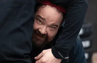 Man peering out from between dark-clad legs, wearing a red cap and beard, with a wary expression