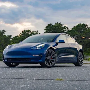Blue Tesla sedan parked on a road, viewed from the front at sunset with trees in the background