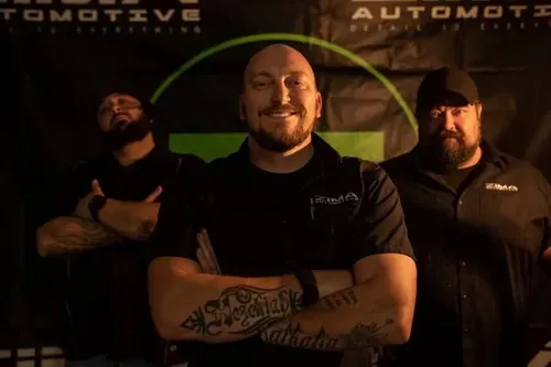 Three tattooed men posing in front of a dark automotive banner, one smiling with arms crossed.
