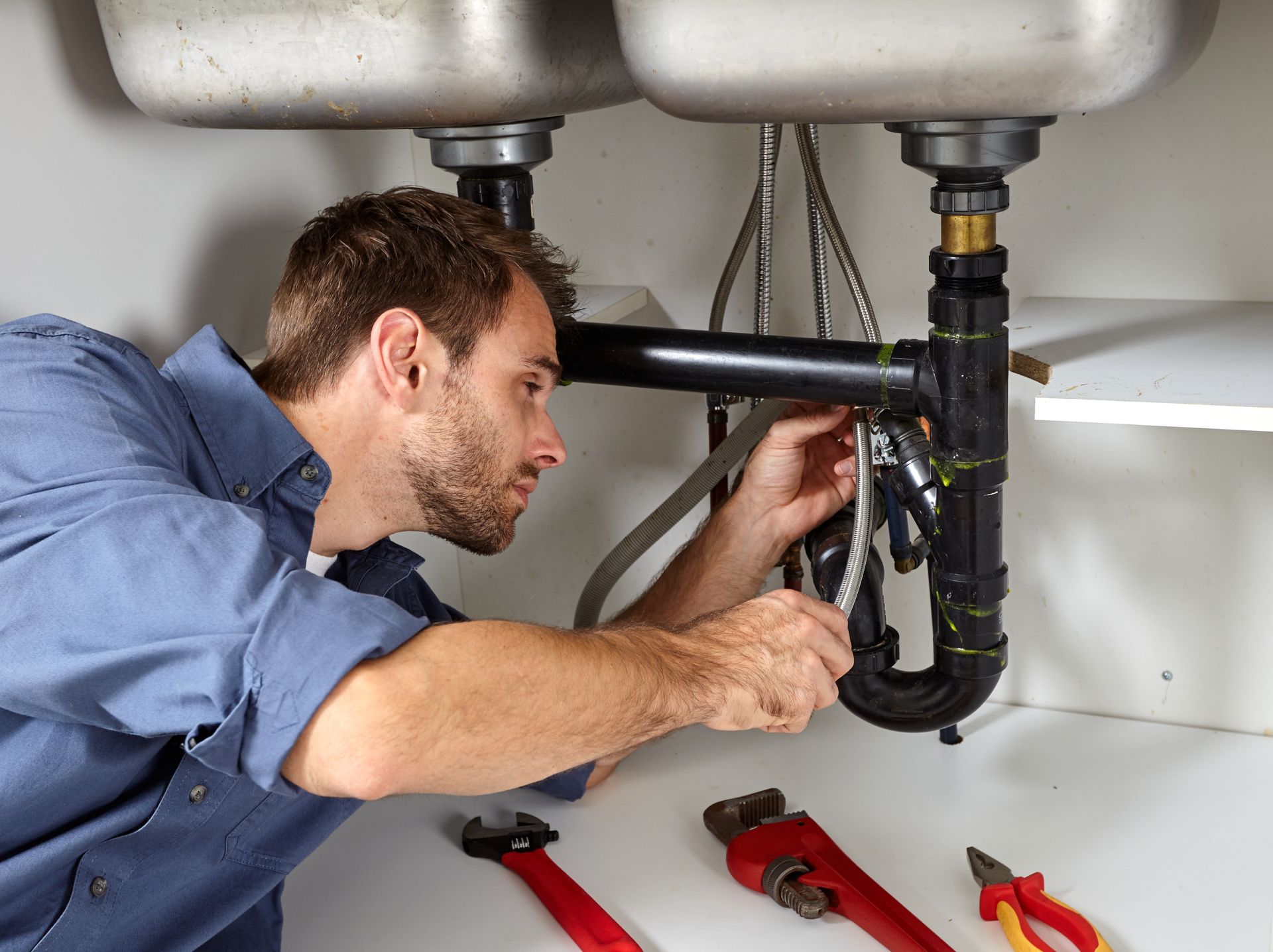 A plumber fixing kitchen sink pipes, wrench in hand, tools scattered on the floor. A plumber fixing kitchen sink pipes, wrench in hand, tools scattered on the floor.