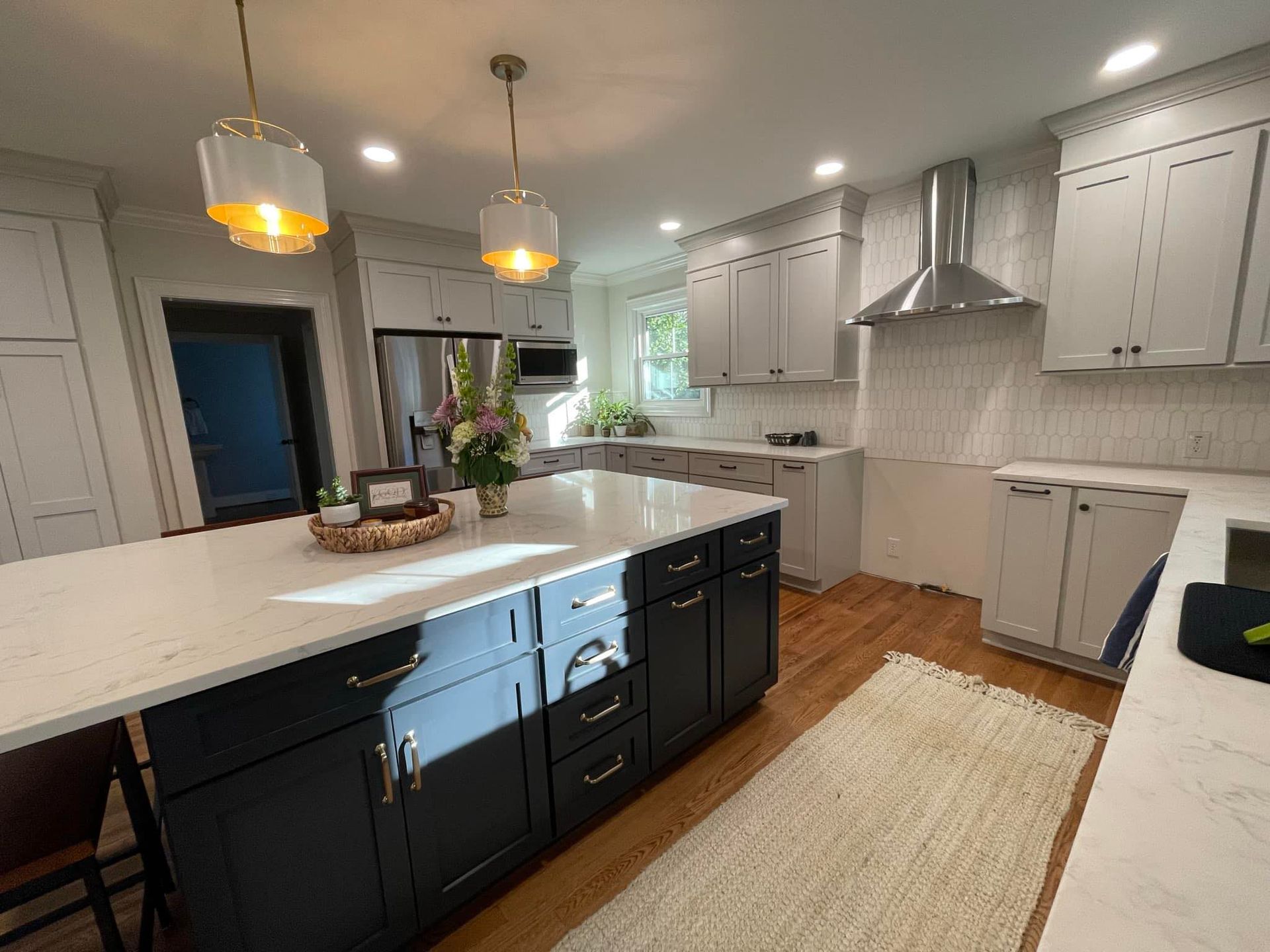 A modern kitchen with a navy island and white cabinets. Wooden floors, a rug, and pendant lights are visible.