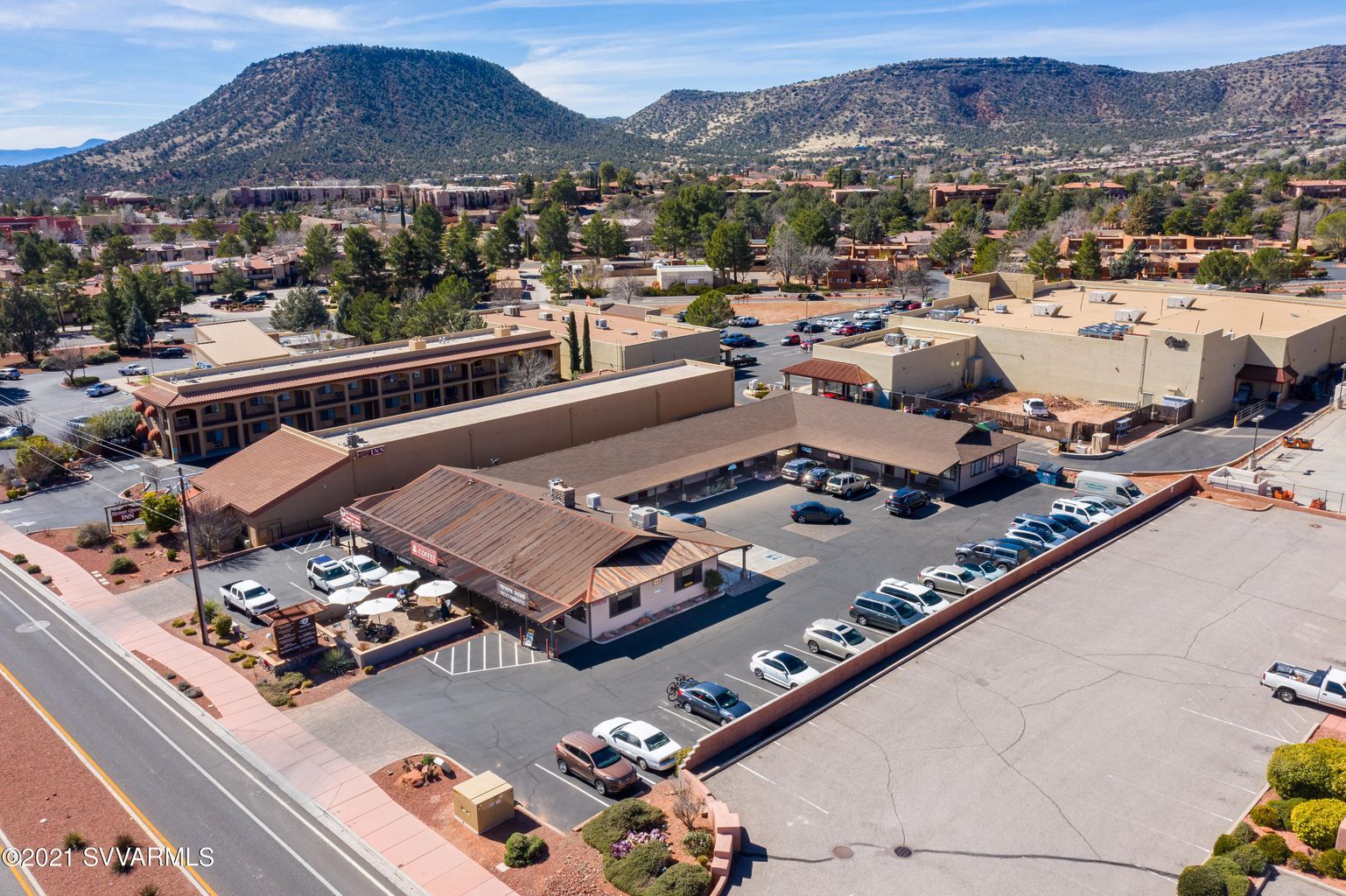 Aerial view of a motel and shops with parking, road, and mountains in the background under a blue sky.