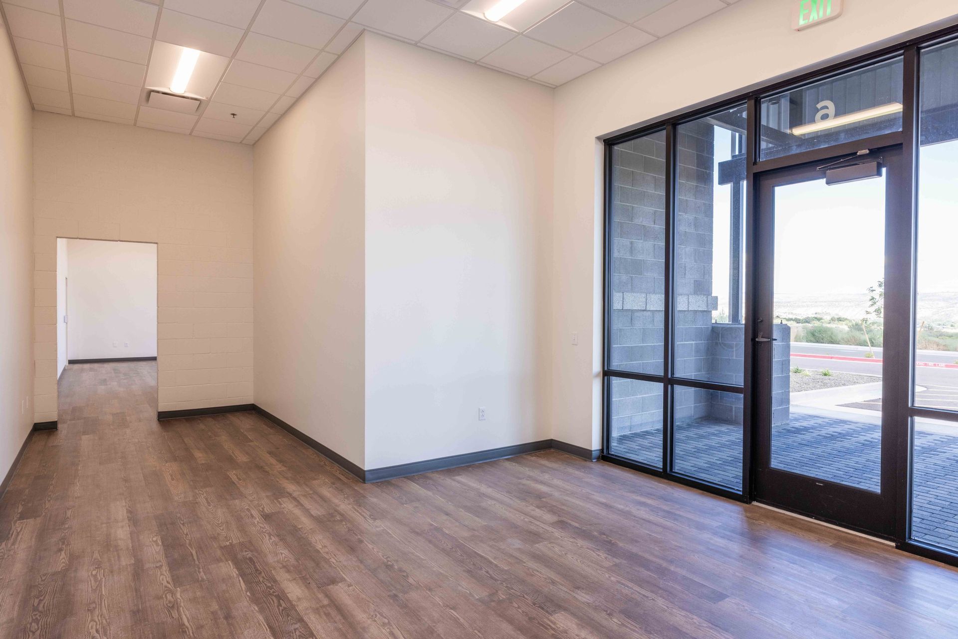 Empty commercial space with wood-look flooring, white walls, and glass door overlooking outdoor view.
