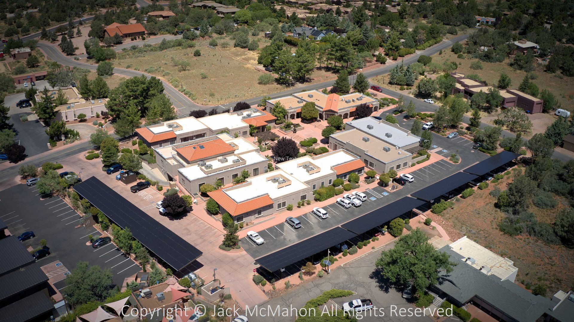 Aerial view of a commercial complex with tan buildings, red-tiled roofs, parking, and trees in a desert landscape.