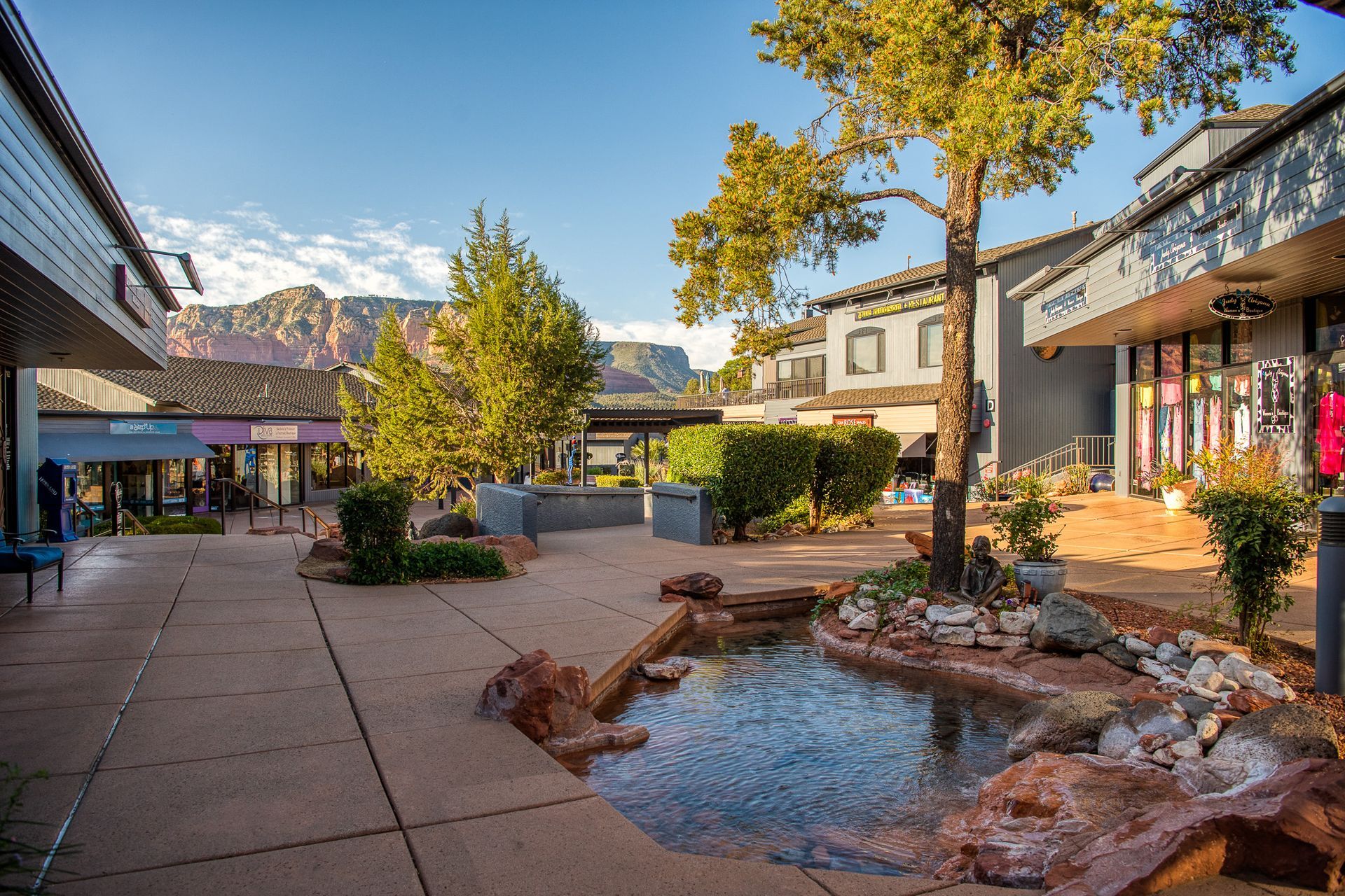Shopping plaza with stores, trees, and small pond against a backdrop of red rock mountains.