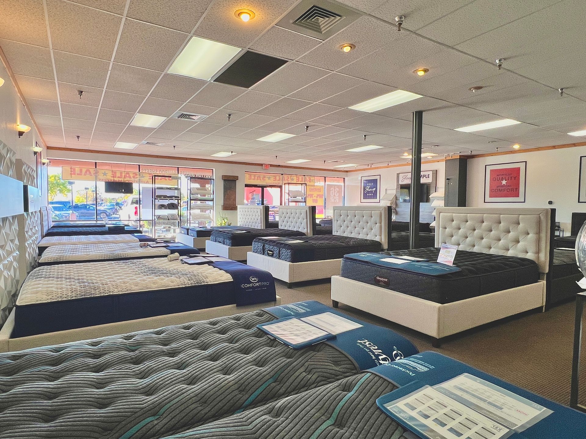 Mattress store interior with various beds on display. Bright lighting, neutral colors, and a view to the outside.