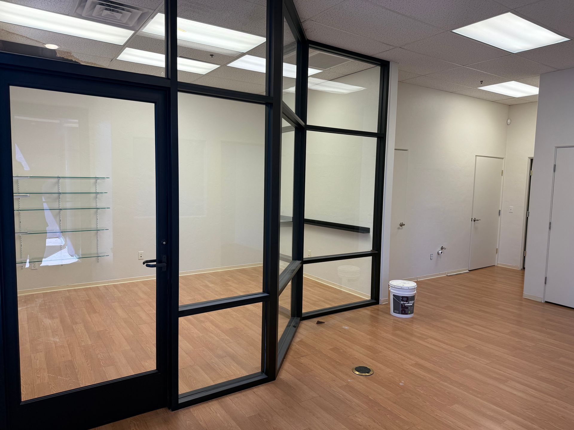 Empty commercial space with glass door and black framing, hardwood floors, and overhead lighting.