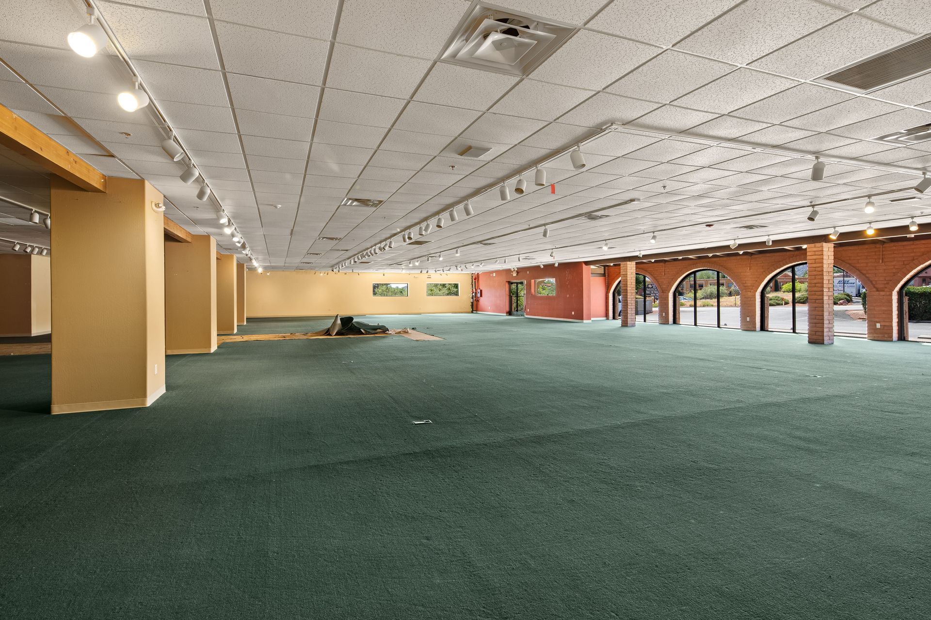 Empty, open-space interior with green carpet, white ceiling, and beige support pillars. Arched doorway leads to exterior.