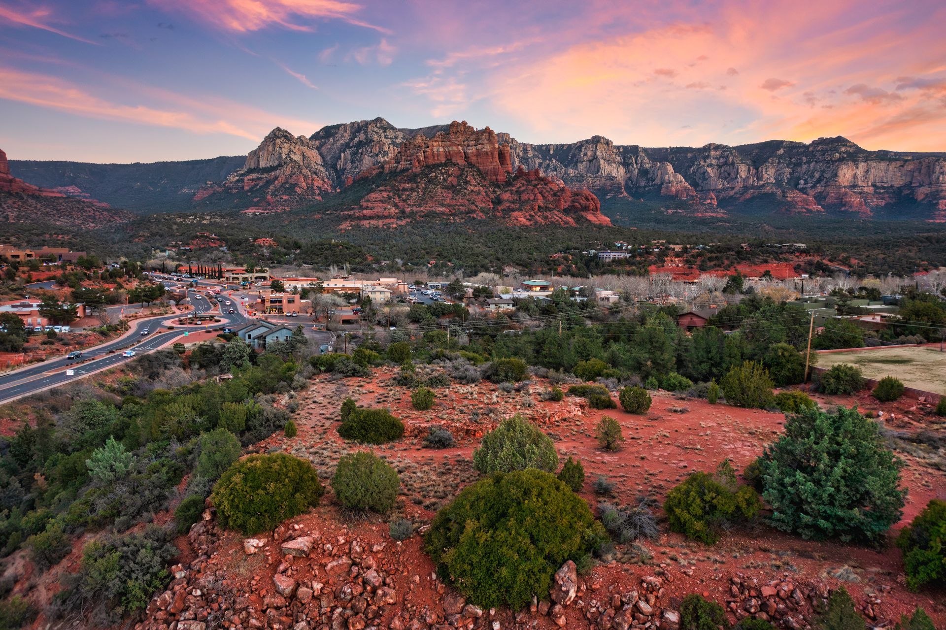 Red rock buttes overlook a town and road. Lush green brush in foreground, pink and blue sunset in sky.