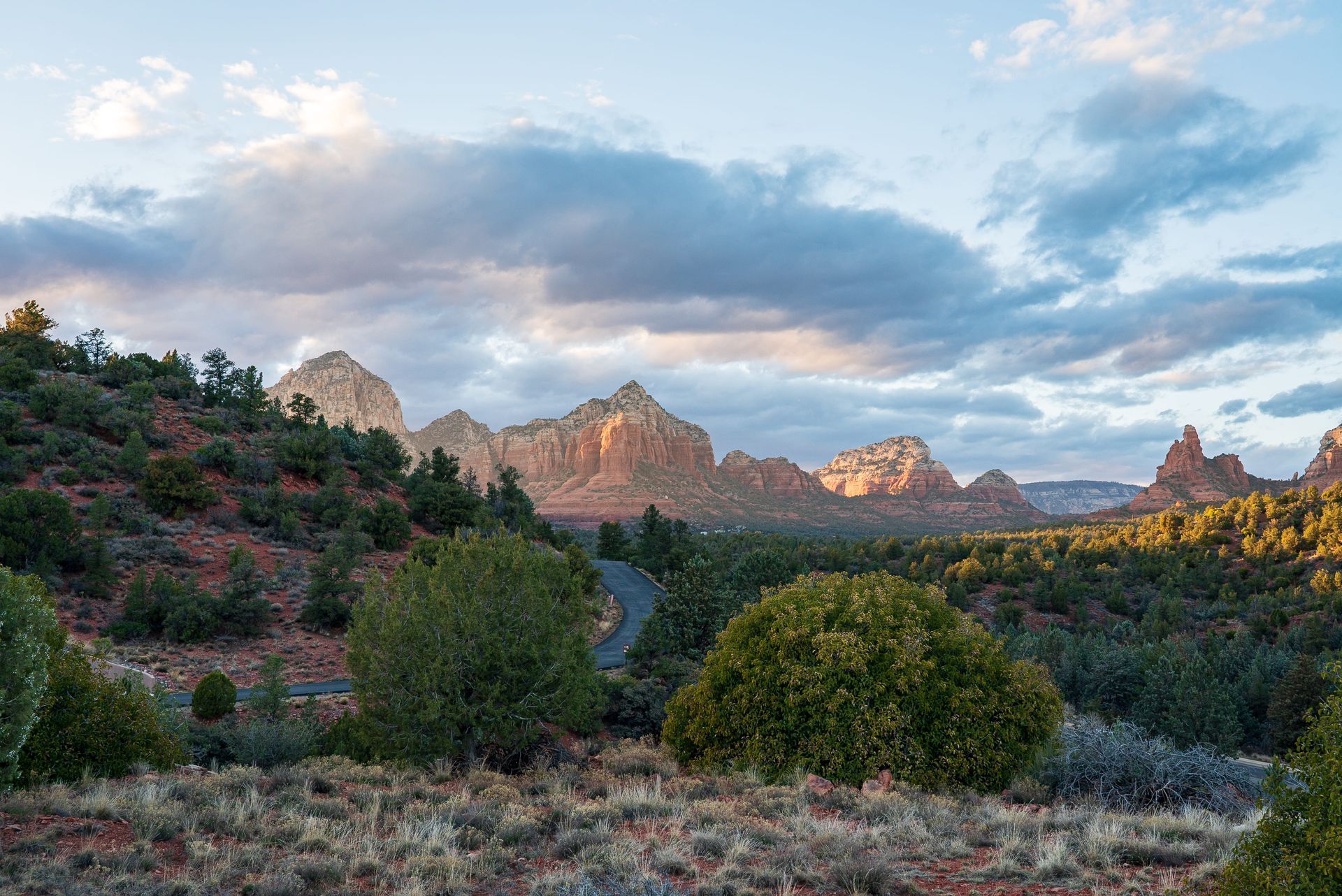 Scenic landscape with red rock formations, green shrubs, and a winding road under a cloudy sky.