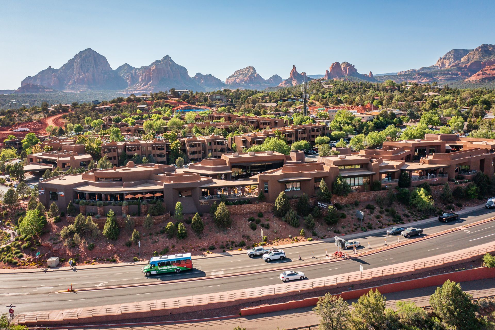 Red-roofed buildings on a hillside in Sedona, Arizona, with red rock formations in the background and cars on a road.