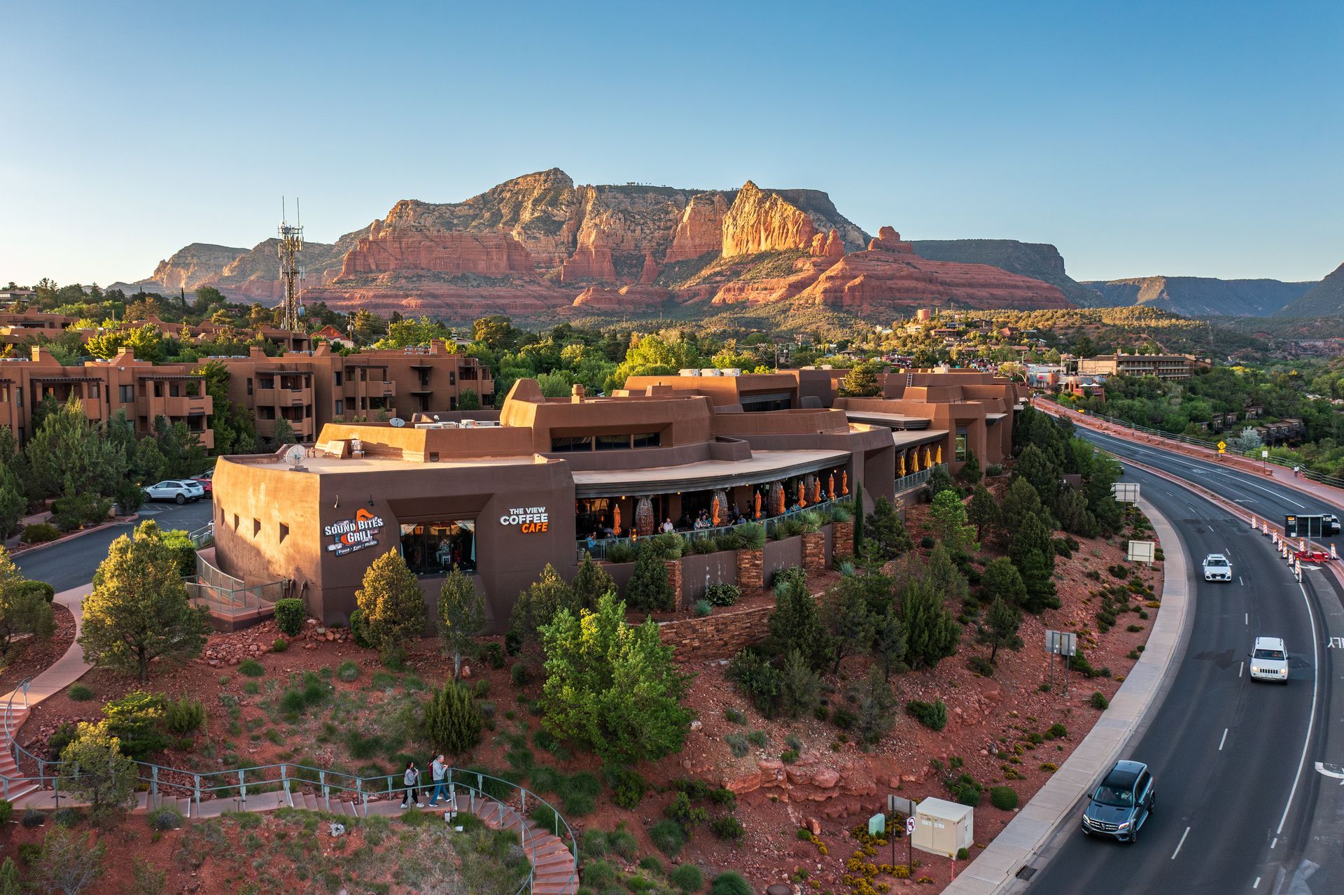 Building with red rock backdrop in Sedona, Arizona. Road and cars in foreground. Trees and blue sky.