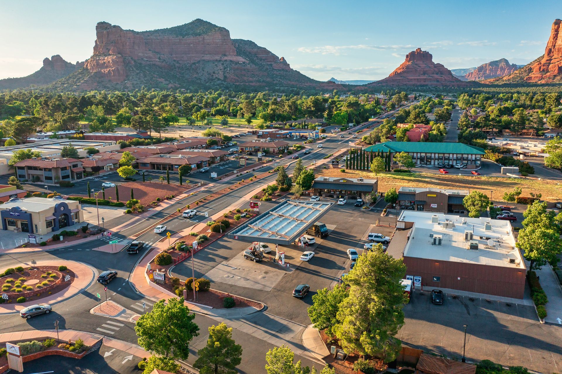 Aerial view of Sedona, Arizona, with red rock formations, a town with buildings, trees, and cars on roads under a blue sky.