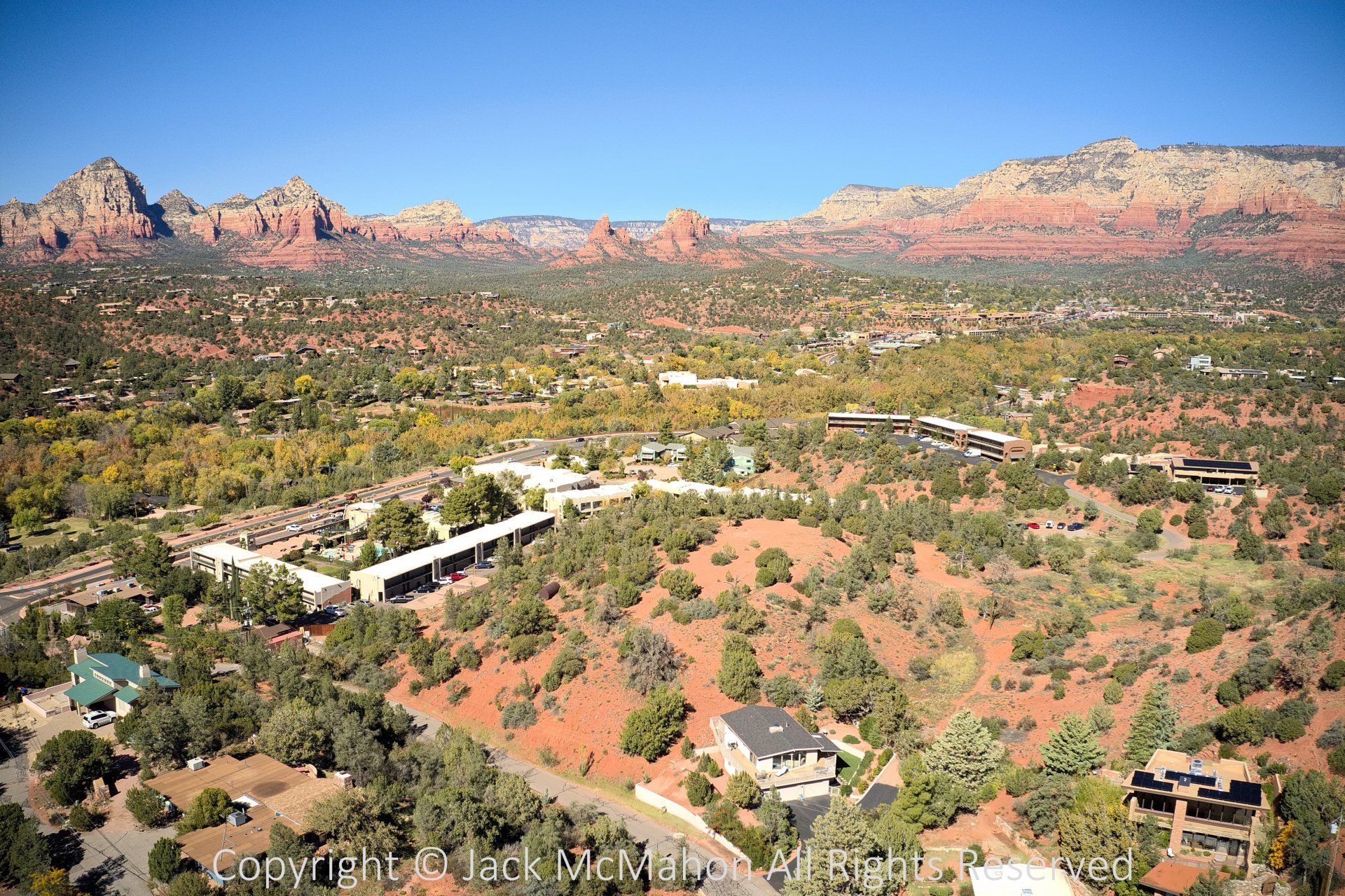 Aerial view of Sedona, Arizona with red rock formations, green trees, and buildings under a blue sky.